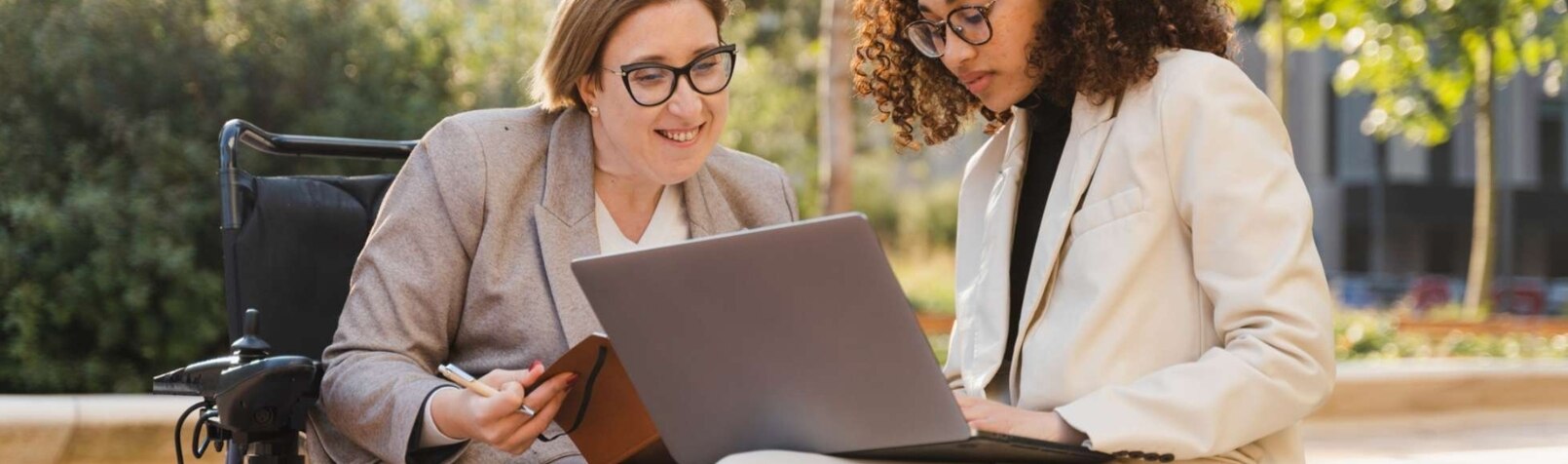 Eine behinderte Frau im Rollstuhl und ihr Geschäftskollege sitzen auf einer Parkbank und arbeiten an einem Laptop, diskutieren und planen ein neues Projekt.