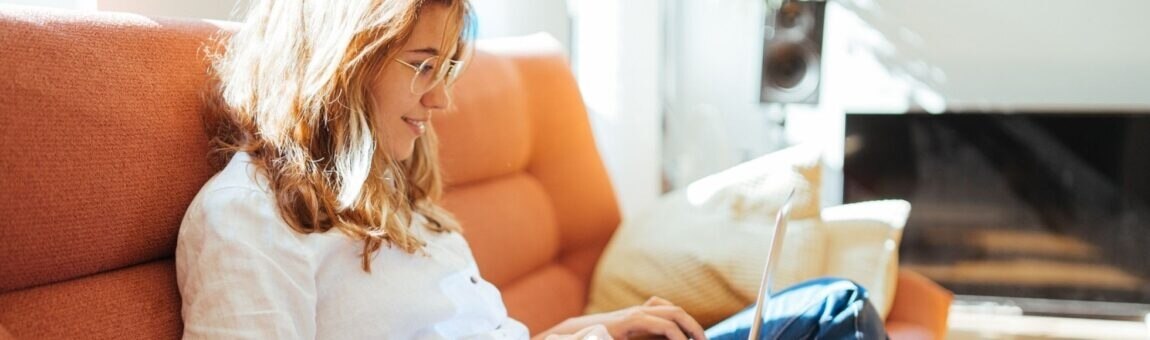 Junge Frau sitzt im Schneidersitz auf dem Sofa und tippt in ihren Laptop.