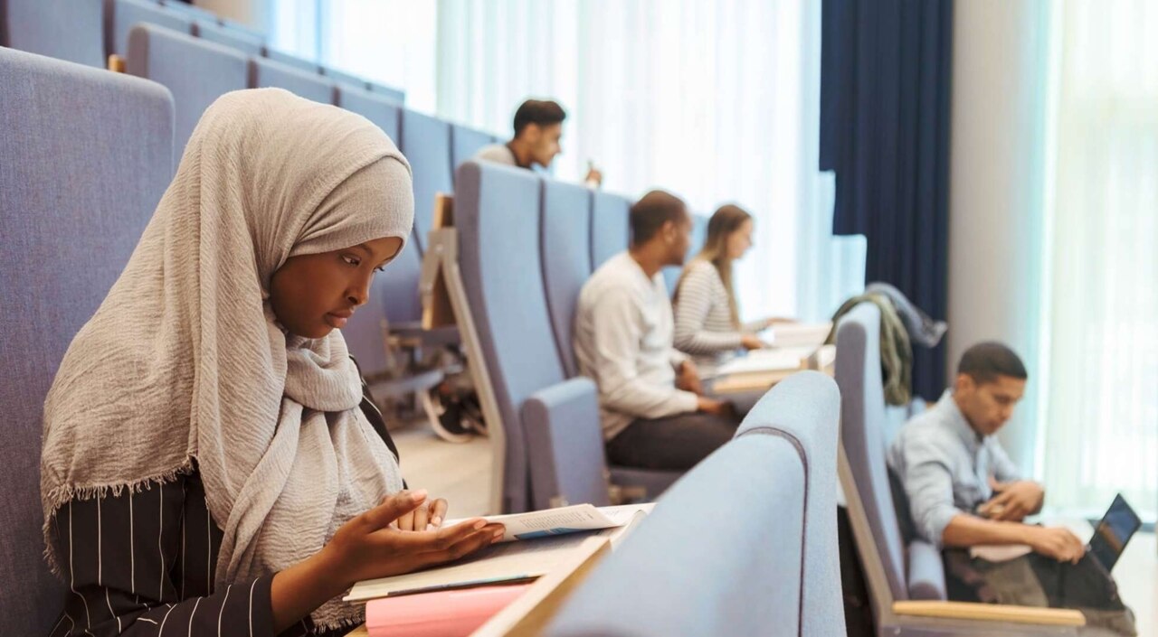 Frau mit Kopftuch sitzt mit anderen Studierenden in einem Hörsaal.