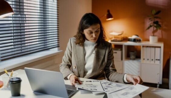 Frau im Business-Outfit arbeitet mit Dokumenten vor einem Laptop im Büro.