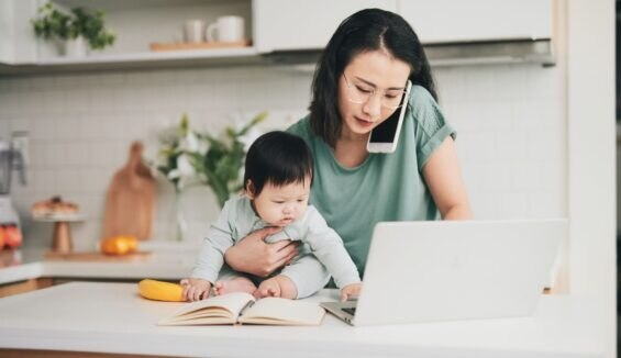 Mutter mit Baby im Arm am Küchentisch, Handy am Ohr und Laptop vor sich auf dem Tisch.