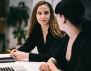 Zwei Frauen sitzen nebeneinander an einem Schreibtisch und schauen sich an