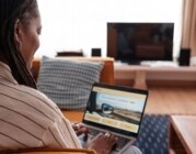 Frau sitzt auf Wohnzimmercouch mit Laptop auf dem Schoß und plant ihren Urlaub.
