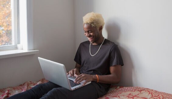 Junger Mann in Jeans und T-Shirt sitzt lächelnd mit seinem Laptop auf seinem Bett.