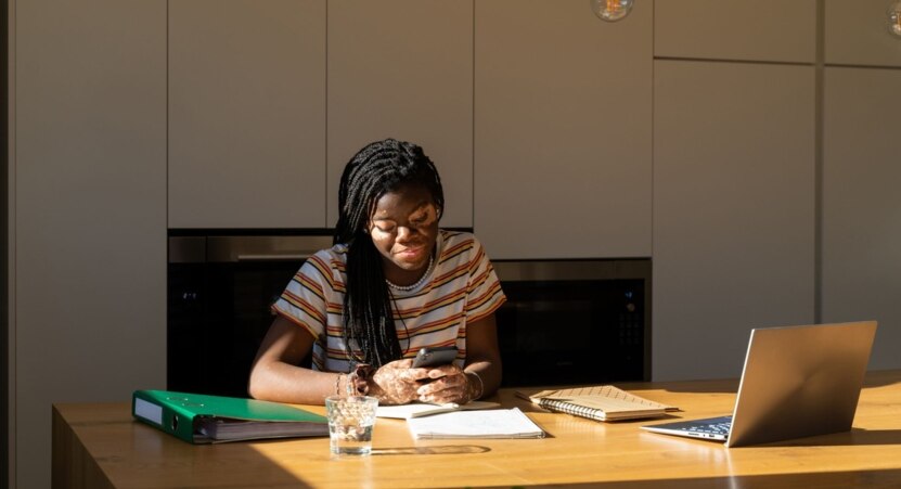 Junge Frau sitzt am Schreibtisch und studiert, blickt auf ihr Handy, vor ihr auf dem Schreibtisch offener Laptop und Schreibmaterial.